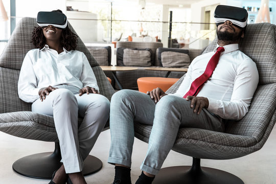 Excited diverse colleagues in VR glasses watching virtual presentation. Business man and woman wearing virtual reality headset, sitting in motion chairs. VR content concept