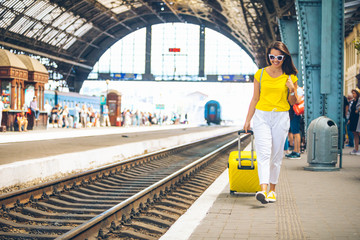 smiling woman walking by railway station with yellow bag on wheels