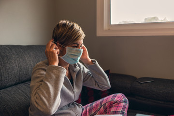 Obraz premium woman in voluntary confinement wearing pajamas putting on surgical masks for the prevention of coronavirus covid virus19. She works at home using the sofa as a desk with a laptop and a smartphone.