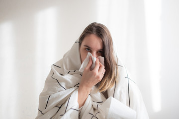 Ill woman under white blanket blowing her nose on white background. Illness treatment, home isolation, quarantine. Copy space