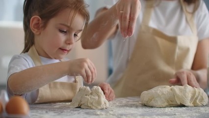 Mother teaching daughter to knead dough for cookies on kitchen in slow motion. Mom teaching kid daughter learning kneading dough with rolling pin, funny child girl helping mother preparing cookies. - Powered by Adobe