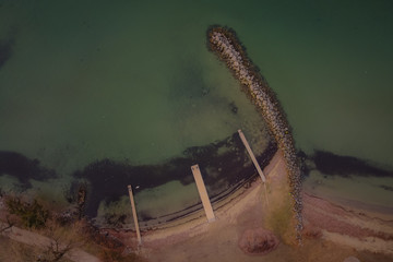 Three small piers viewed directly from above shielded by long stone barrier in water. Small empty marina seen from above, green water.