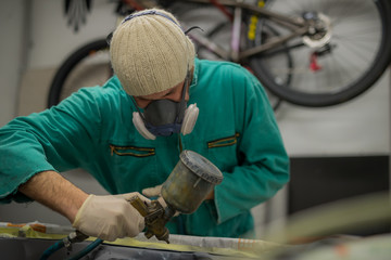 Man in green overalls preparing an engine bay of a car for a final coat of paint. Man spraying grey liquid putty or stucco on the car panels as a last layer before final paint coat.