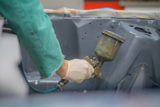 Man in green overalls preparing an engine bay of a car for a final coat of paint. Man spraying grey liquid putty or stucco on the car panels as a last layer before final paint coat.