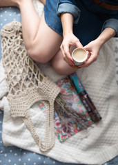 Seated woman on a woolen blanket besides a crochet bag and a lifestyle magazine.
