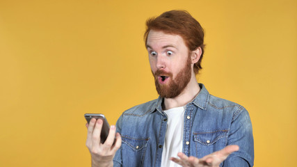 Amazed Redhead Man Surprised while Using Smartphone Isolated on Yellow Background