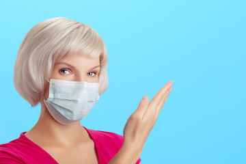 A beautiful girl with blond hair in a protective medical surgical mask looks at the camera and points with her hand to the empty space for text. Guidelines for prevention or protection against disease