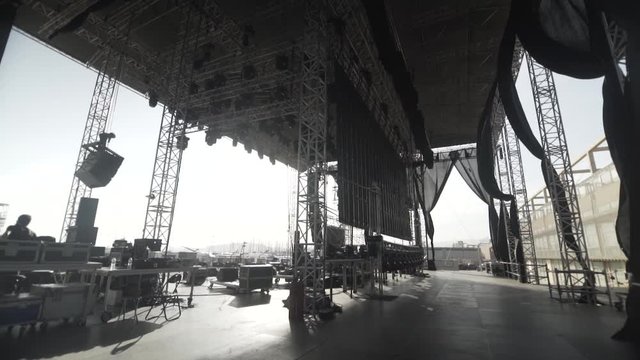 day shot of an enormous music stage before a live concert metallic and black elements , black fabrics slowly moves from the wind , flight cases on stage