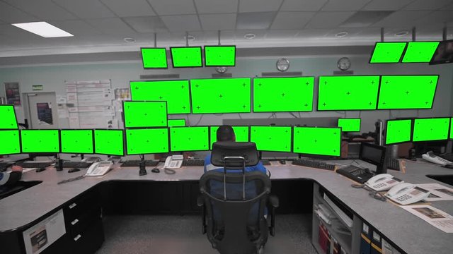 A worker sits in front of many monitors. Green screen with markers. Production room.