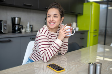 Fototapeta na wymiar Young woman sitting in the kitchen, holding cup, smiling