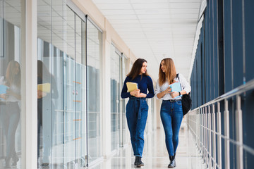 Two young female students standing with books and bags in the hallway University speaking each other.