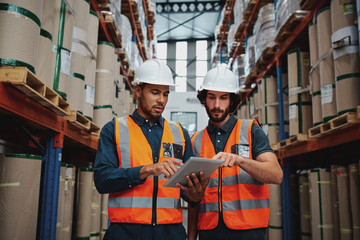 Focused manager and employee standing in warehouse discussing inventory stock while standing between cardboard packed goods with safety hardhat and vest