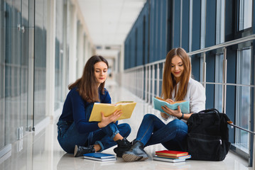 two pretty female students with books sitting on the floor in the university hallway