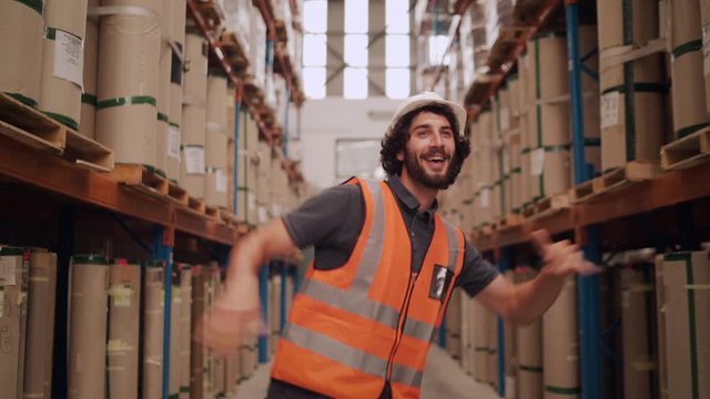 A caucasian young male worker in uniform dancing and having fun between palettes with ordered goods and materials at warehouse