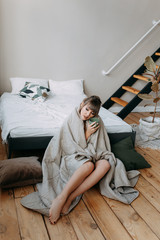 Beautiful happy sweet lonely girl woke up and wrapped in a blanket sitting on the bed in a spacious bright bedroom, resting and drinking coffee, holidays, weekends, hospital regime, cozy home