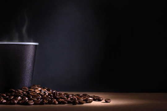 Black disposable cup of coffee with smoke and scattered coffee beans on dark background. Space for text