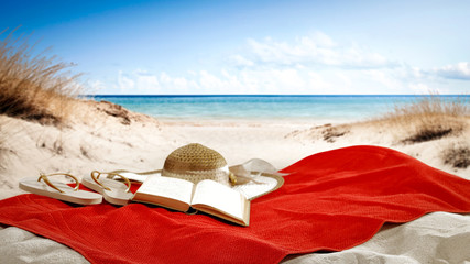 Red towel on beach and book with summer hat. 