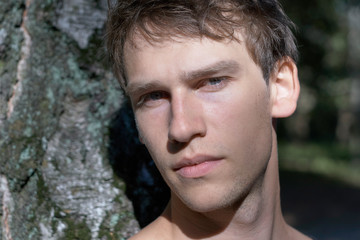 Portrait of an unshaven handsome young man leaning against a tree and looking serious into the camera.