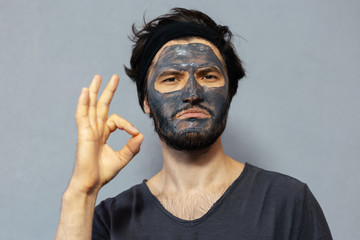 Portrait of young disheveled guy with facial mask showing OK gesture with hand on background of grey background.
