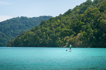 Fototapeta na wymiar A Person is playing a standing surfing board on the blue turquoise sea of Andaman ocean - Thailand. Outdoor sport and recreation action photo.