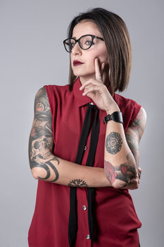 Bella ragazza mora con una camicia smanicata rossa e una cravatta nera, piena di tattuaiggi nelle braccia &egrave; dubbiosa , isolata su sfondo grigio