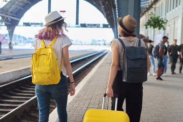 Two traveling women with suitcase backpacks walking along platform