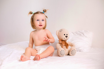 Toddler girl in diapers sits with toy bear on bed at home