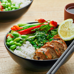 Wholesome meal made of teriyaki chicken fillet, fresh cucumber, red bell pepper, edamame, stew cabbage and basmati rice. Two delicious Buddha bowls with green tea cups on a table.