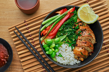 Wholesome meal made of teriyaki chicken fillet, fresh cucumber, red pepper, edamame, stew cabbage and rice. Delicious Buddha bowl and green tea cup on a table. Top view, directly above.