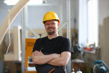 Portrait of young attractive man in work clothes and yellow helmet crossed arms smiling at shop for manufacturing furniture and details home interior