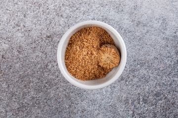 Nutmeg seed and ground nutmeg on concrete background. Freshly grated spice in white bowl. Close-up, copy space, top view.