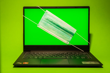 Laptop on a green background with a surgical mask on the monitor.  The computer shows a green background on the display.