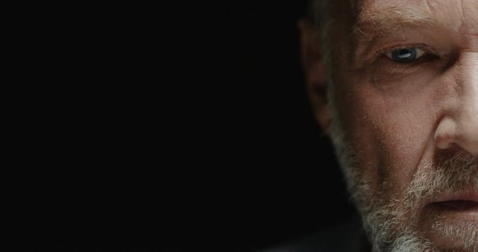 Close up portrait of good-looking Caucasian senior grey-haired man half face on black background looking with serious expression indoors. Handsome bearded old male posing to camera in light shadow