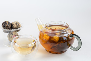 Blooming black tea in a glass teapot. On a light surface. Nearby are tea briquettes of leaves and flowers. In the background is a cake.