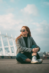 Stylish young beautiful girl in fashionable spring clothes with white sneakers sits on the asphalt