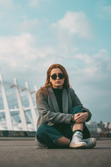Stylish young beautiful girl in fashionable spring clothes with white sneakers sits on the asphalt