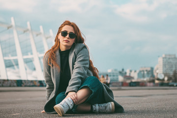 Stylish young beautiful girl in fashionable spring clothes with white sneakers sits on the asphalt
