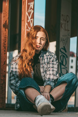 Stylish young beautiful girl in fashionable spring clothes with white sneakers sits on the asphalt