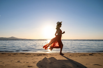 Young woman wearing long red dress and straw hat running on sand beach at sea shore enjoying view...
