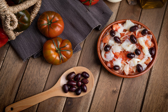 Esqueixada in clay pot and wooden spoon with some black olives on black napkin. zenith view