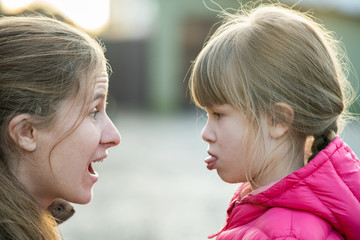 Close up portrait of young mom and her daughter girl making faces to each other outdoors.