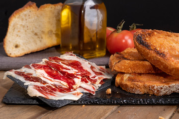 Still life of serrano ham with tomato bread on slate stone tray. black background.