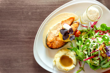 Grilled salmon steak sliced on white plate with mixed vegetable salad, mashed potatos and topping in a glass, food in dish on wooden table, top view and copy space