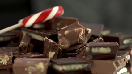 Close up view on rotating pieces of chocolate on the table with black and white background with red and white candy or lollipop on top. A lot of different chunks of chocolate.