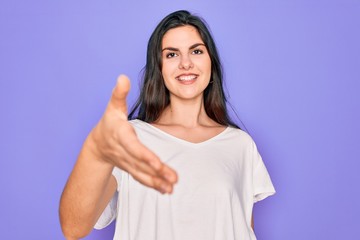 Young beautiful brunette woman wearing casual white t-shirt over purple background smiling friendly offering handshake as greeting and welcoming. Successful business.