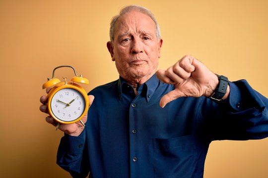 Senior grey haired man holding vintage alarm clock over yellow background with angry face, negative sign showing dislike with thumbs down, rejection concept
