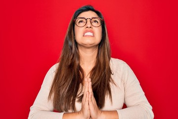 Young hispanic smart woman wearing glasses standing over red isolated background begging and...