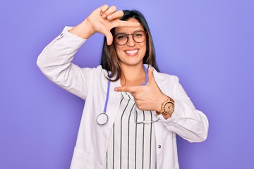 Professional doctor woman wearing stethoscope and medical coat over purple background smiling making frame with hands and fingers with happy face. Creativity and photography concept.