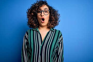 Young beautiful curly arab woman wearing striped shirt and glasses over blue background afraid and...