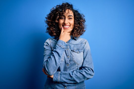 Young beautiful curly arab woman wearing casual denim shirt standing over blue background looking confident at the camera smiling with crossed arms and hand raised on chin. Thinking positive.